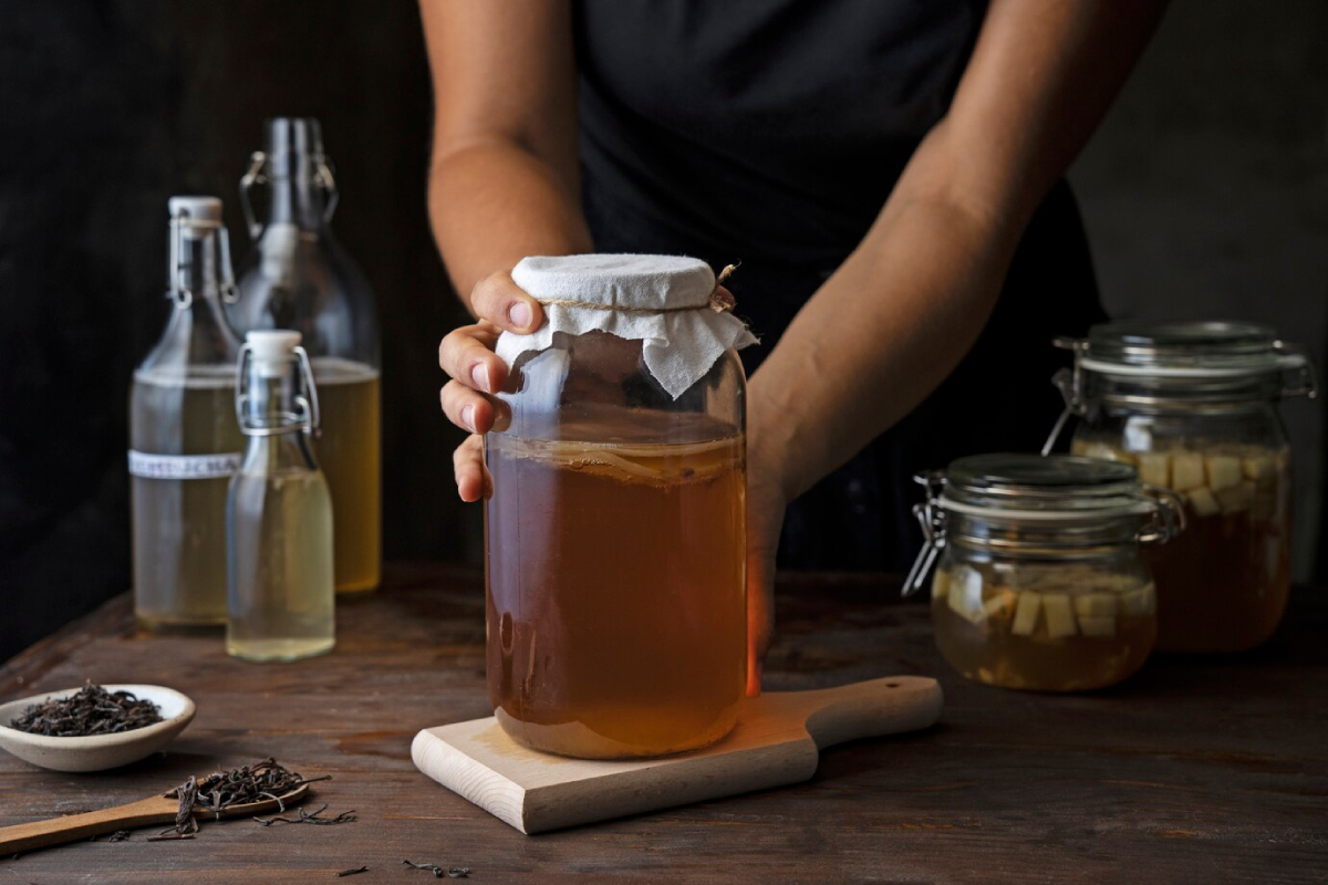 A person holding a fermentation jar of kombucha, surrounded by various jars and bottles, with loose tea leaves in the foreground.