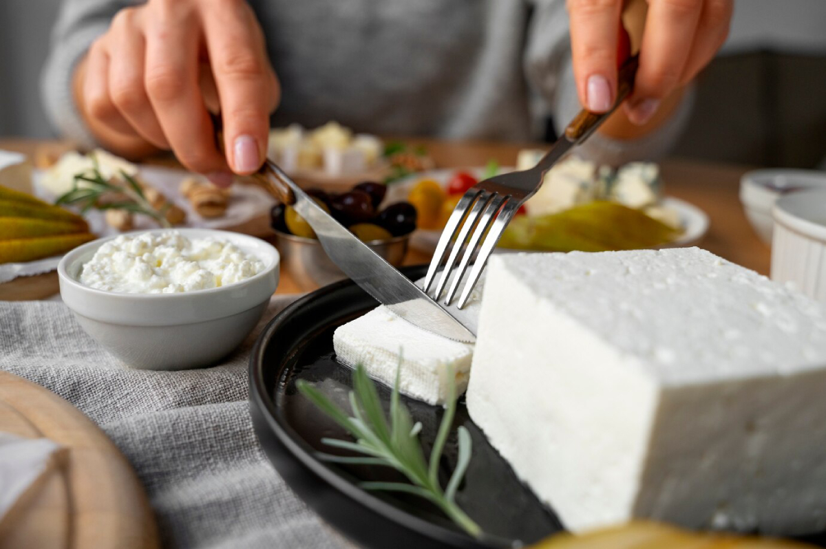 A close-up of hands slicing creamy cheese on a black plate, surrounded by olives, pickles, and a bowl of cottage cheese.