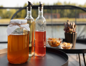 A jar of amber liquid with a cloth cover, flanked by glass bottles and bowls of snacks on a black table by a lake.