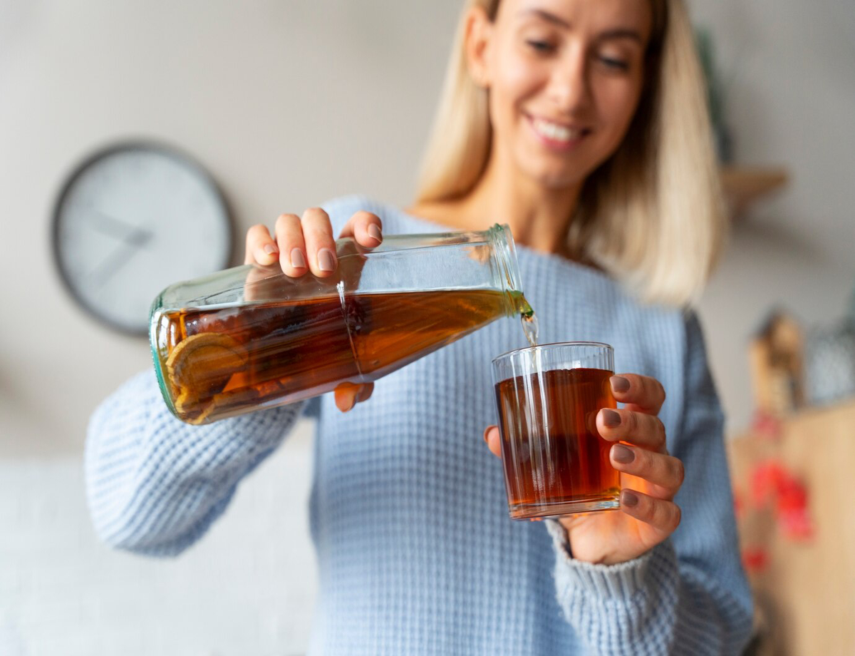 A person in a cozy sweater pours amber liquid from a glass bottle into a clear glass on a wooden table, with a clock in the background.