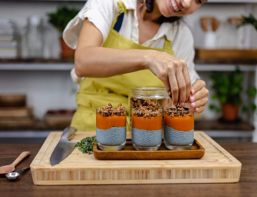 A woman in a yellow apron decorates chia seed desserts with granola, smiling while standing in a cozy kitchen filled with plants.
