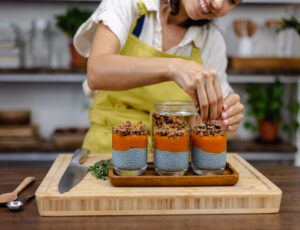 A woman in a yellow apron decorates chia seed desserts with granola, smiling while standing in a cozy kitchen filled with plants.