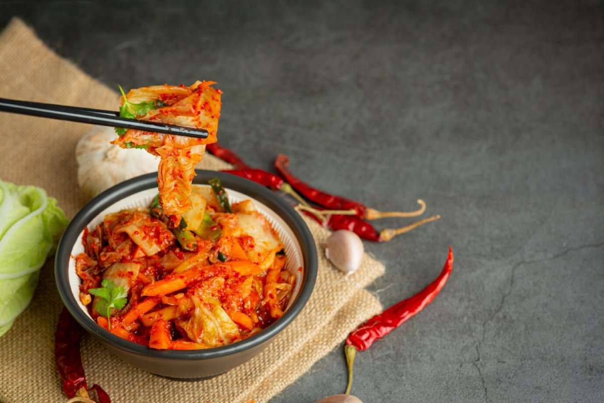 A bowl of vibrant kimchi with chopsticks picking up a piece, surrounded by garlic, red chilies, and lettuce on a burlap background.