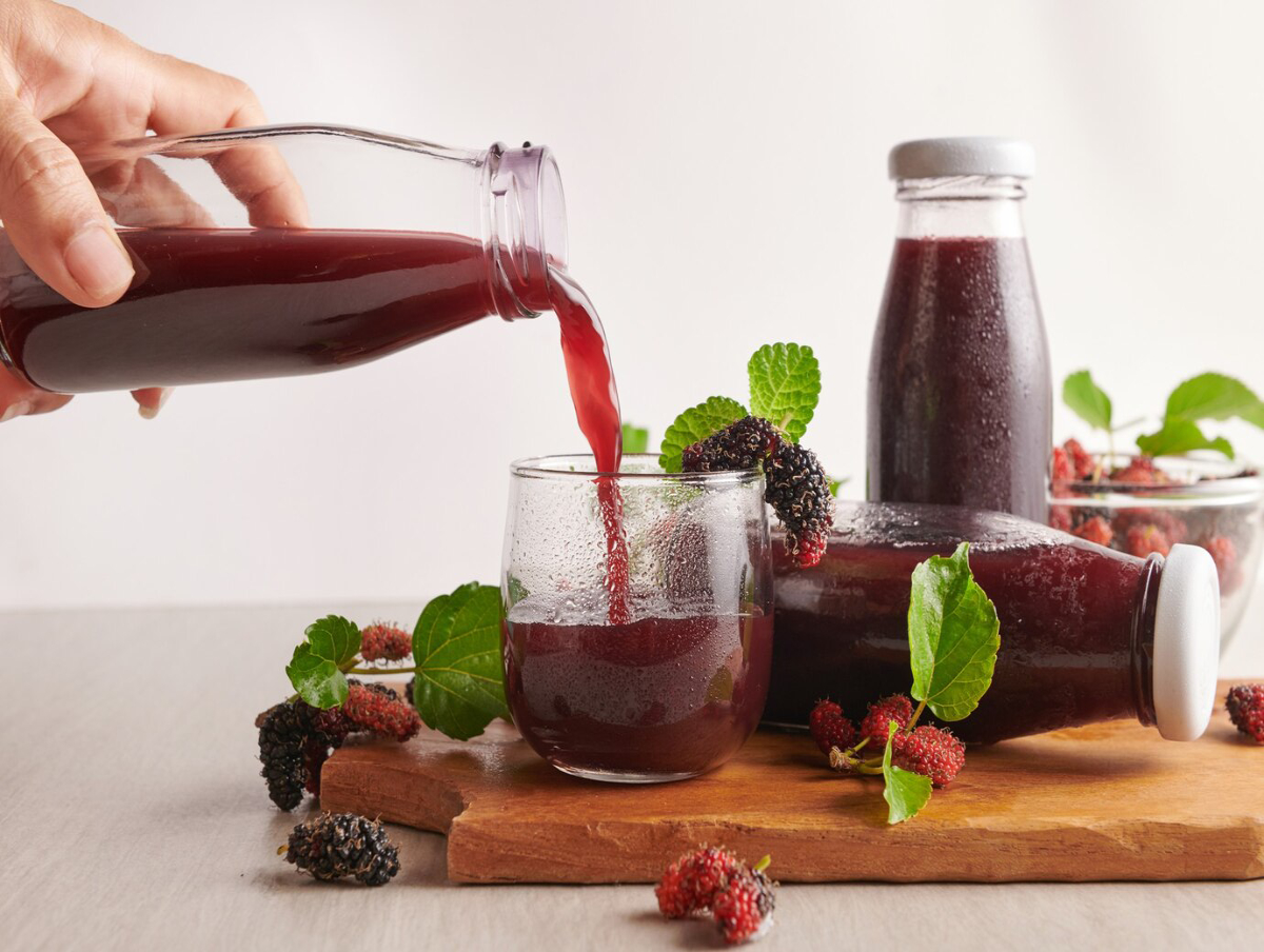 A hand pours dark red juice from a glass bottle into a chilled glass, surrounded by fresh mulberries and mint leaves on a wooden board.