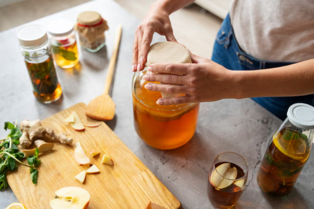 Hands cover a large jar of kombucha on a countertop. Nearby are apple slices, ginger, herbs, a wooden spoon, and jars filled with liquid. Inviting, natural tone.
