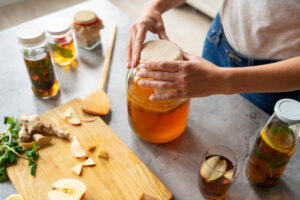 Hands cover a large jar of kombucha on a countertop. Nearby are apple slices, ginger, herbs, a wooden spoon, and jars filled with liquid. Inviting, natural tone.