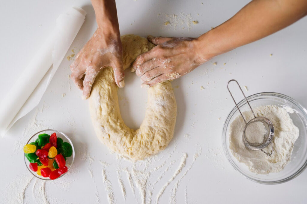 A pair of hands knead dough on a white surface, surrounded by flour, a rolling pin, and a bowl of colorful gummy candies.