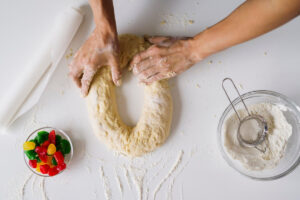 A pair of hands knead dough on a white surface, surrounded by flour, a rolling pin, and a bowl of colorful gummy candies.
