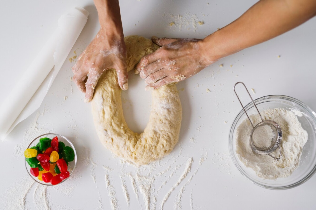 A pair of hands knead dough on a white surface, surrounded by flour, a rolling pin, and a bowl of colorful gummy candies.