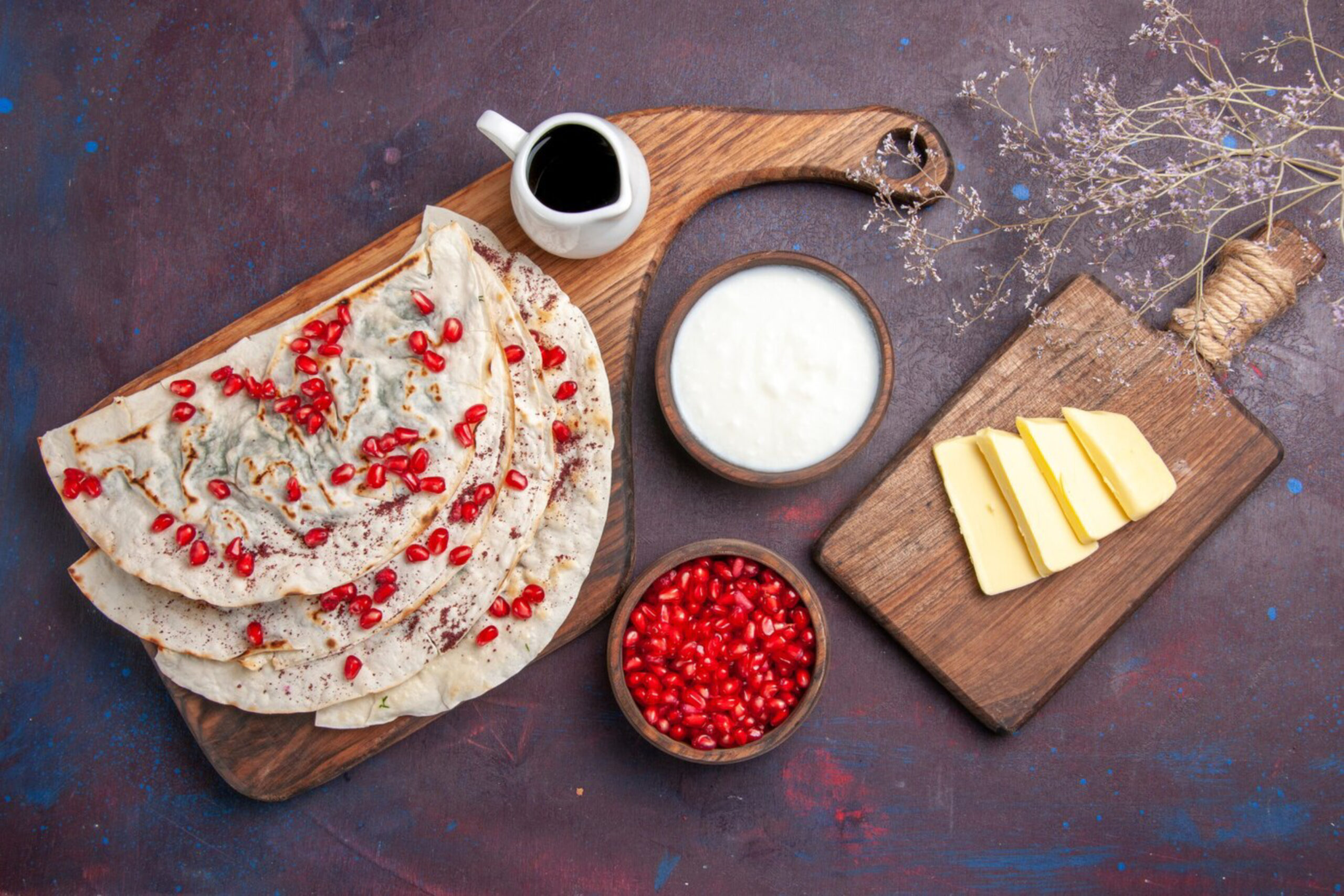 A rustic wooden board with flatbreads topped with pomegranate seeds. Nearby are bowls of yogurt, pomegranate arils, a pitcher of molasses, and sliced cheese.