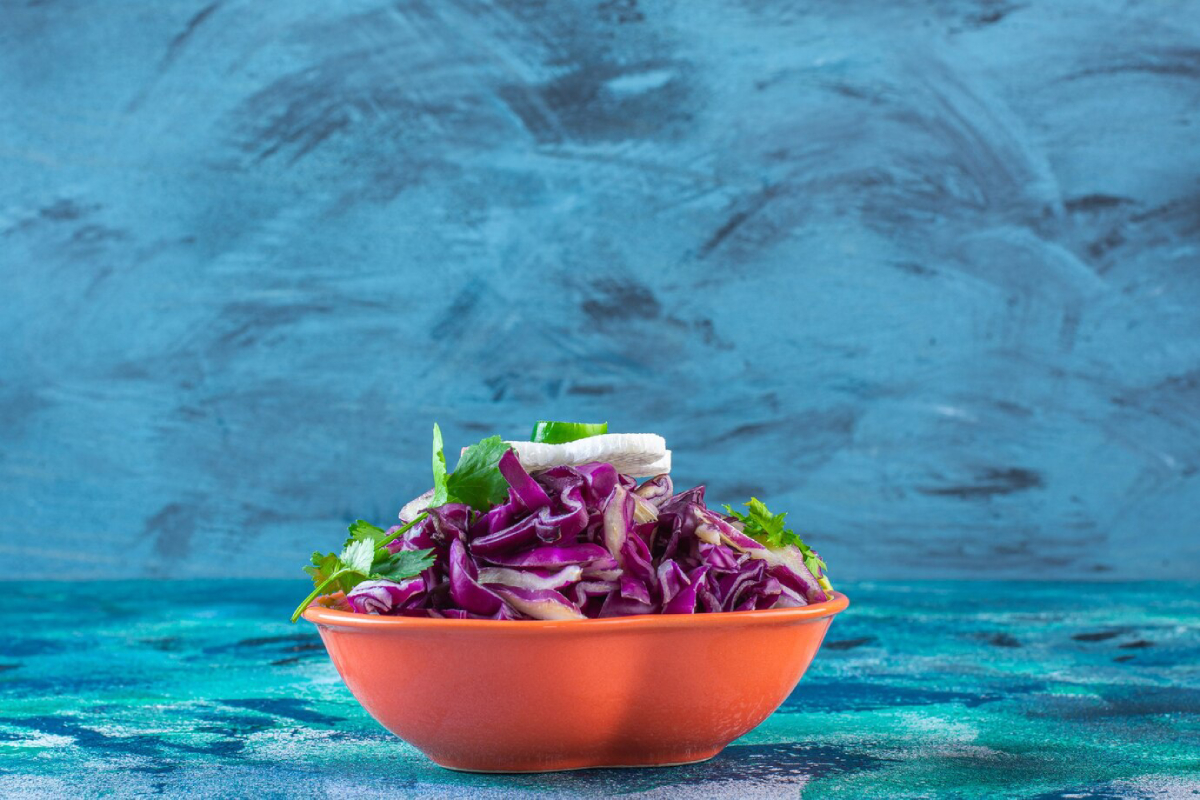 A vibrant orange bowl filled with finely shredded purple cabbage and fresh cilantro, set against a textured blue background.