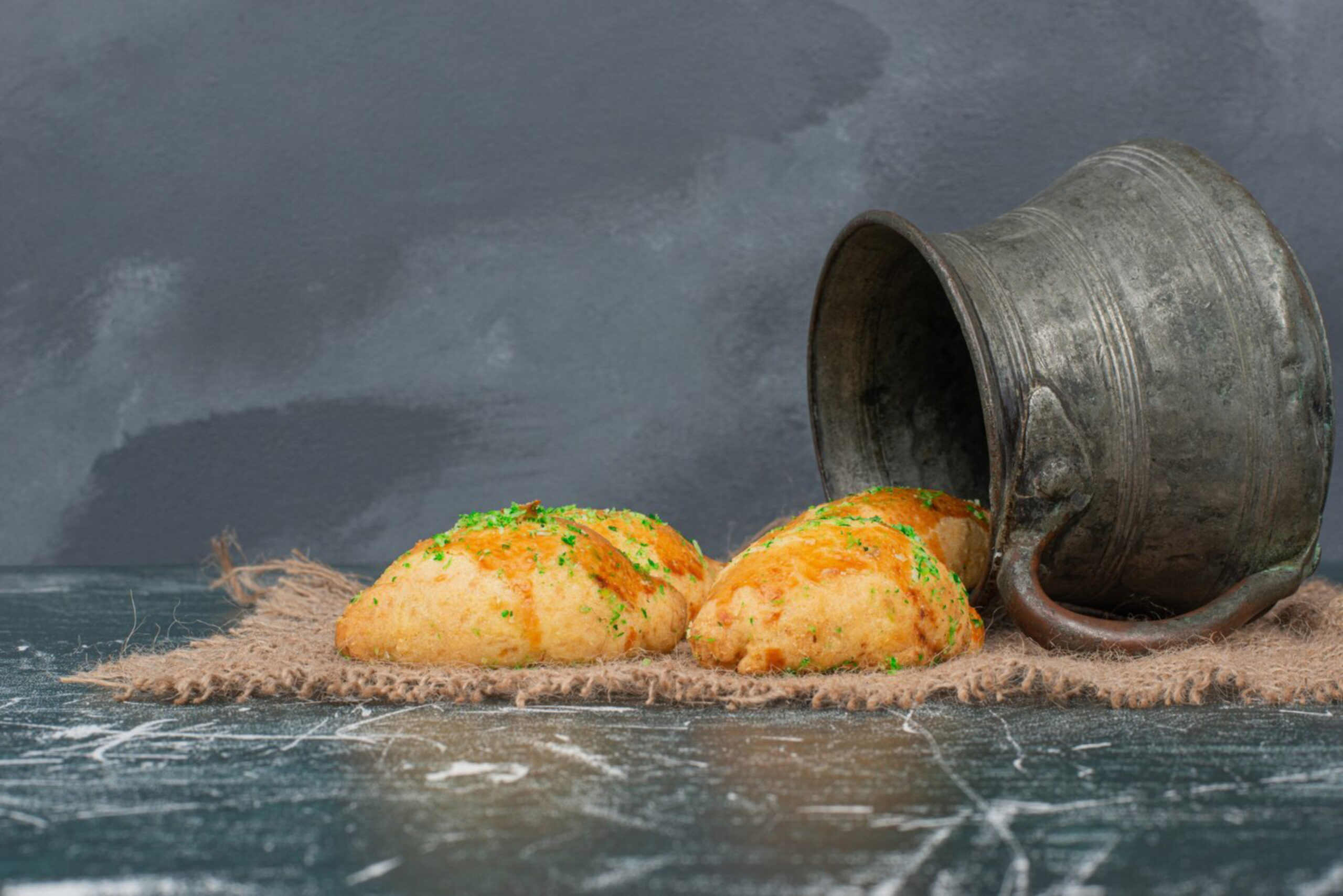 Golden bread rolls garnished with herbs spill from a rustic metal pot onto a burlap cloth against a textured gray background. Cozy and inviting.