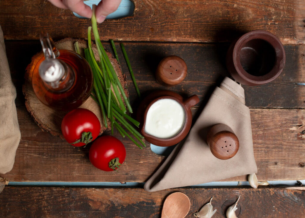 Alt text: Rustic kitchen scene with tomatoes, green onions, vinaigrette, and cream in clay dishes on a wooden table. Hand holds green onions. Cozy, earthy tones.