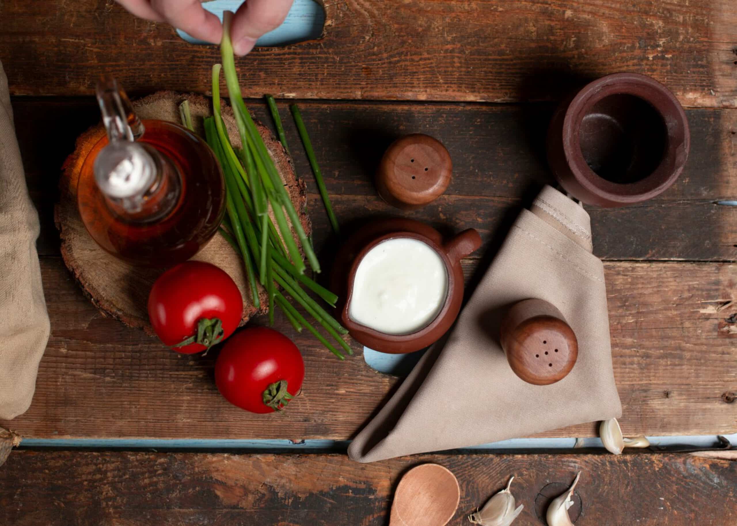 Alt text: Rustic kitchen scene with tomatoes, green onions, vinaigrette, and cream in clay dishes on a wooden table. Hand holds green onions. Cozy, earthy tones.