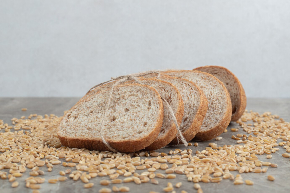 Sliced whole grain bread tied with twine, resting on a surface sprinkled with grains, against a soft gray background.