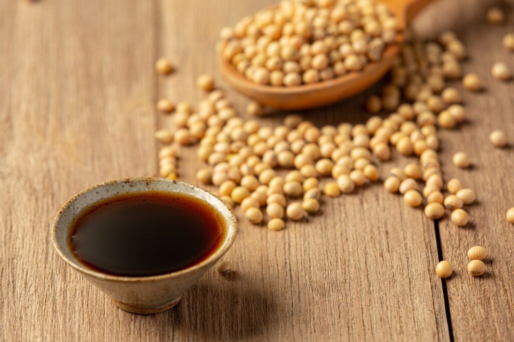 A small ceramic bowl filled with dark soy sauce sits on a wooden surface beside scattered light-colored soybeans and a wooden spoon.
