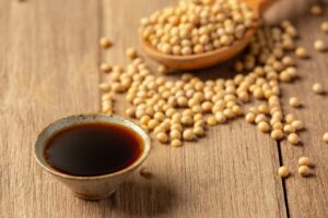 A small ceramic bowl filled with dark soy sauce sits on a wooden surface beside scattered light-colored soybeans and a wooden spoon.