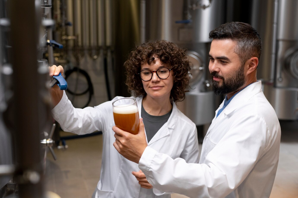 Two individuals in white lab coats are inspecting brewing equipment, one holding a glass of amber beer in a brewery setting.