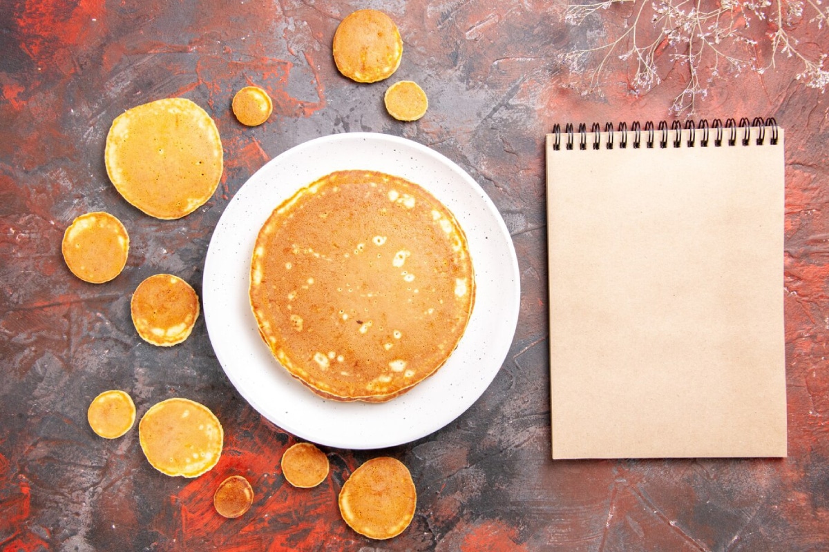 A plate of round pancakes, varying in size, surrounded by smaller pancakes and a blank notepad on a textured dark background.