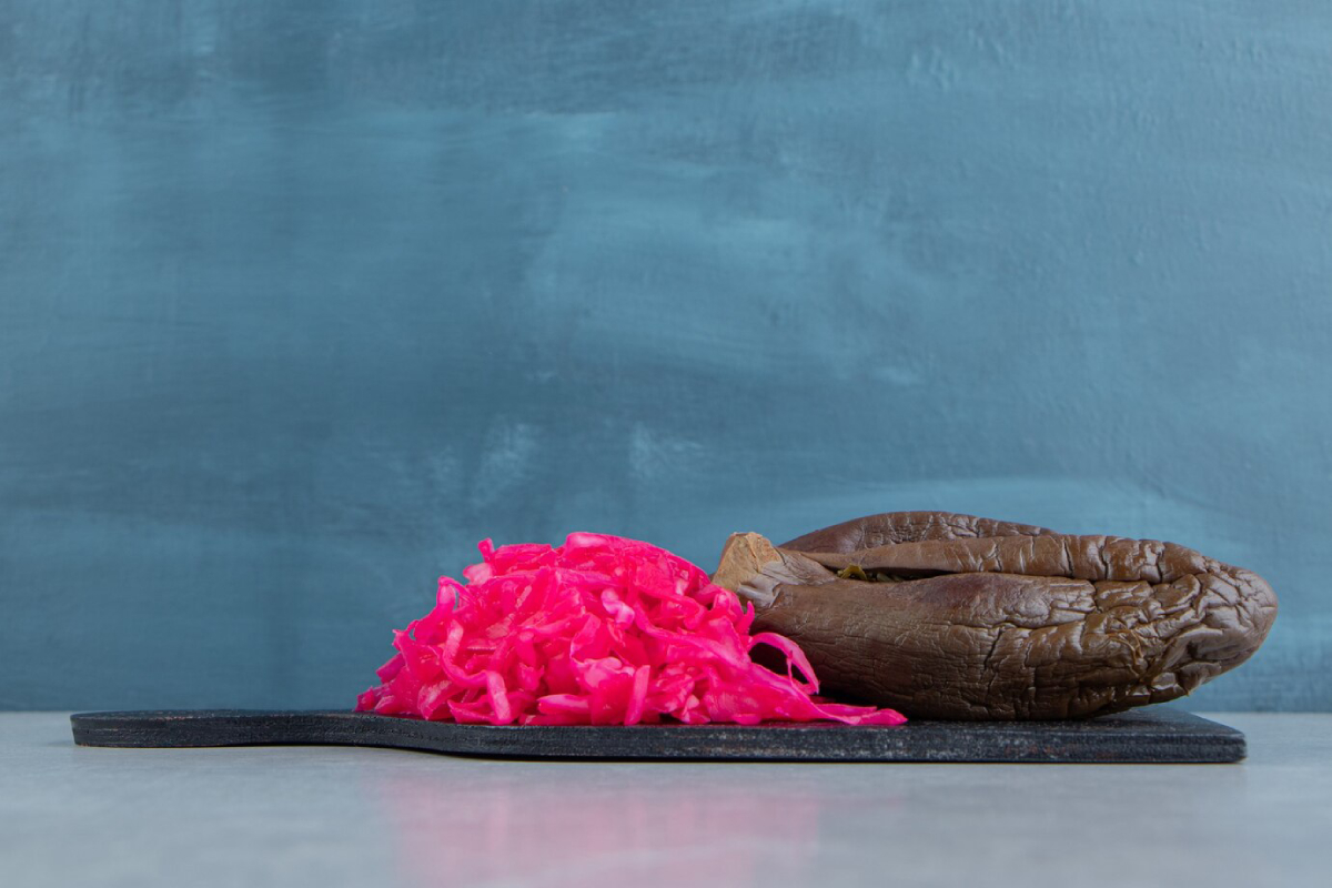 A vibrant pile of pink pickled cabbage next to a dark, wrinkled eggplant on a black board against a soft blue background.