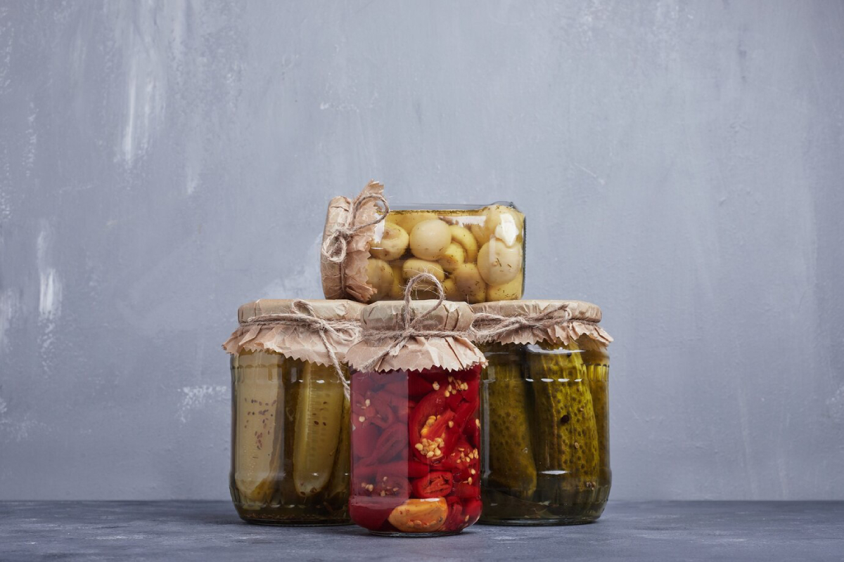 A stack of glass jars filled with pickled vegetables, including cucumbers, peppers, and small pickled onions, tied with rustic twine.