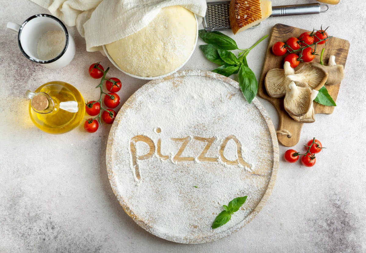 A wooden pizza board dusted with flour, featuring the word "pizza" etched in the flour, surrounded by ingredients like tomatoes, basil, and mushrooms.