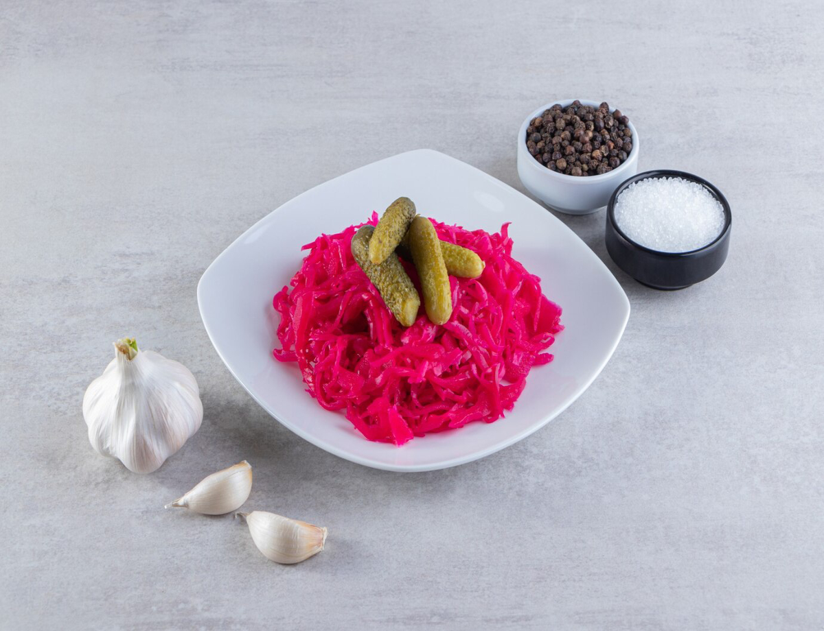 A plate of vibrant pink pickled cabbage topped with pickles, surrounded by garlic cloves and small bowls of pepper and salt.