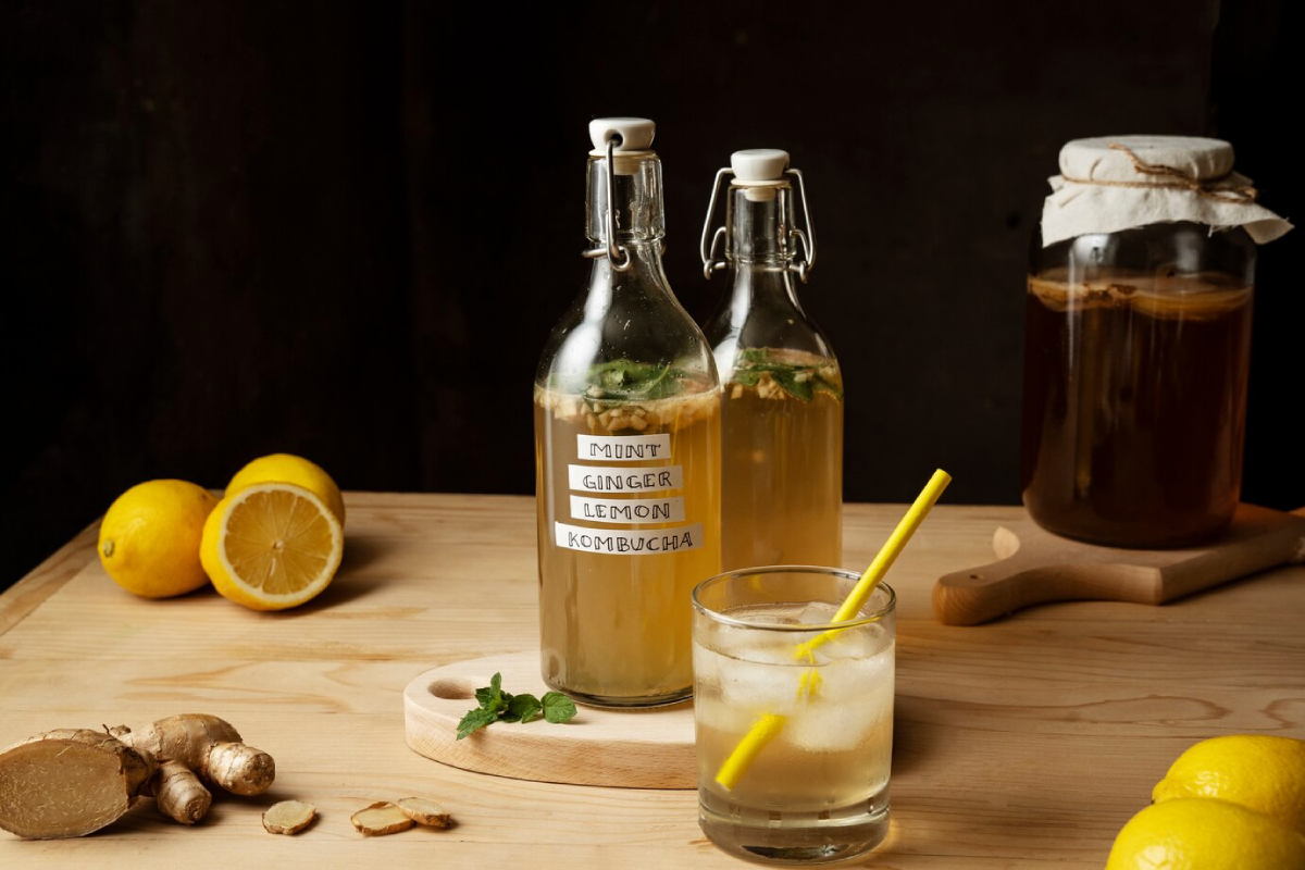 Two glass bottles of mint, ginger, and lemon kombucha, a filled glass with a yellow straw, ginger, and lemons on a wooden table.