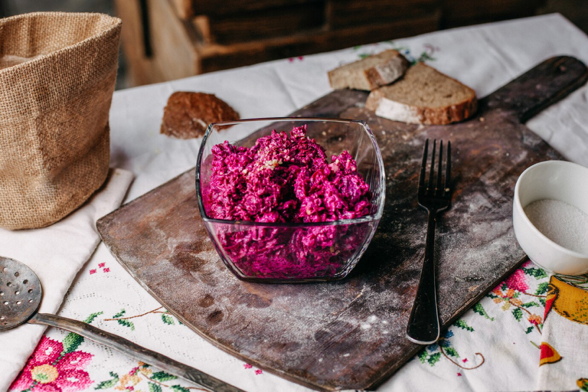 A bowl of vibrant purple beet salad on a wooden cutting board, with bread, a fork, and a sugar bowl nearby.