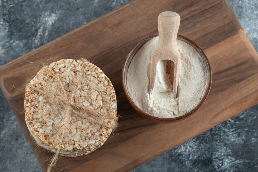 A wooden bowl filled with flour and a scoop, next to a round stack of puffed rice tied with twine on a wooden cutting board.