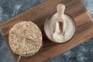 A wooden bowl filled with flour and a scoop, next to a round stack of puffed rice tied with twine on a wooden cutting board.
