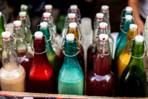 Colorful bottles filled with vibrant liquids, featuring varied shades of red, green, blue, and yellow, sit in a rustic wooden container.