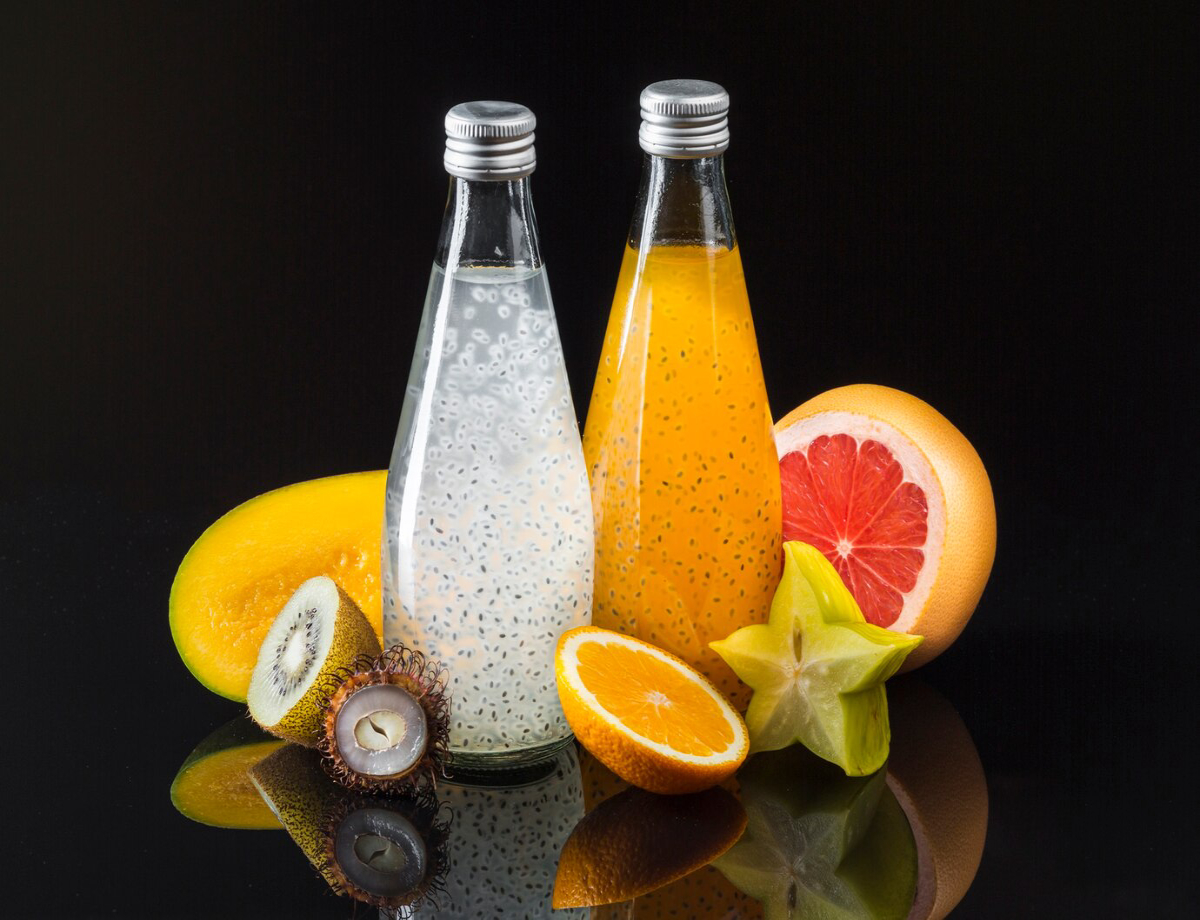 Two glass bottles of fresh fruit juice, one clear with chia seeds and one vibrant orange, surrounded by assorted tropical fruits.
