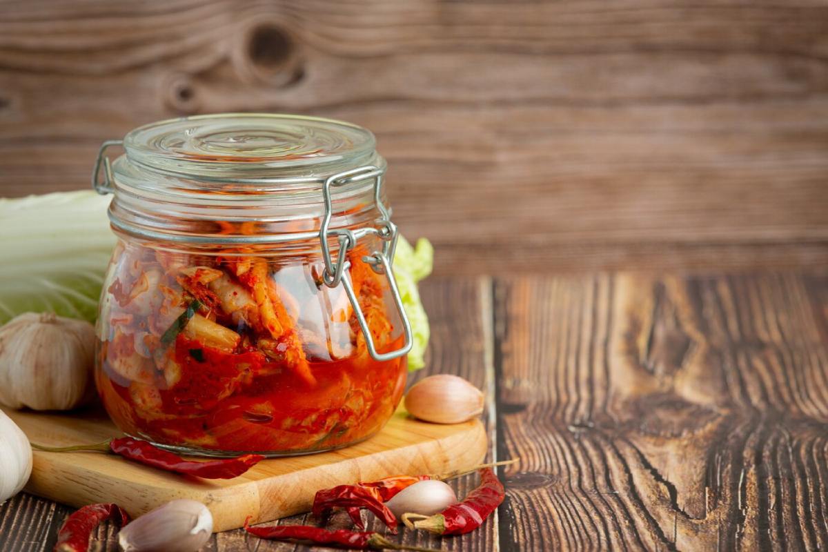 A jar of vibrant kimchi sits on a wooden cutting board, surrounded by garlic, shallots, dried chilies, and fresh cabbage.