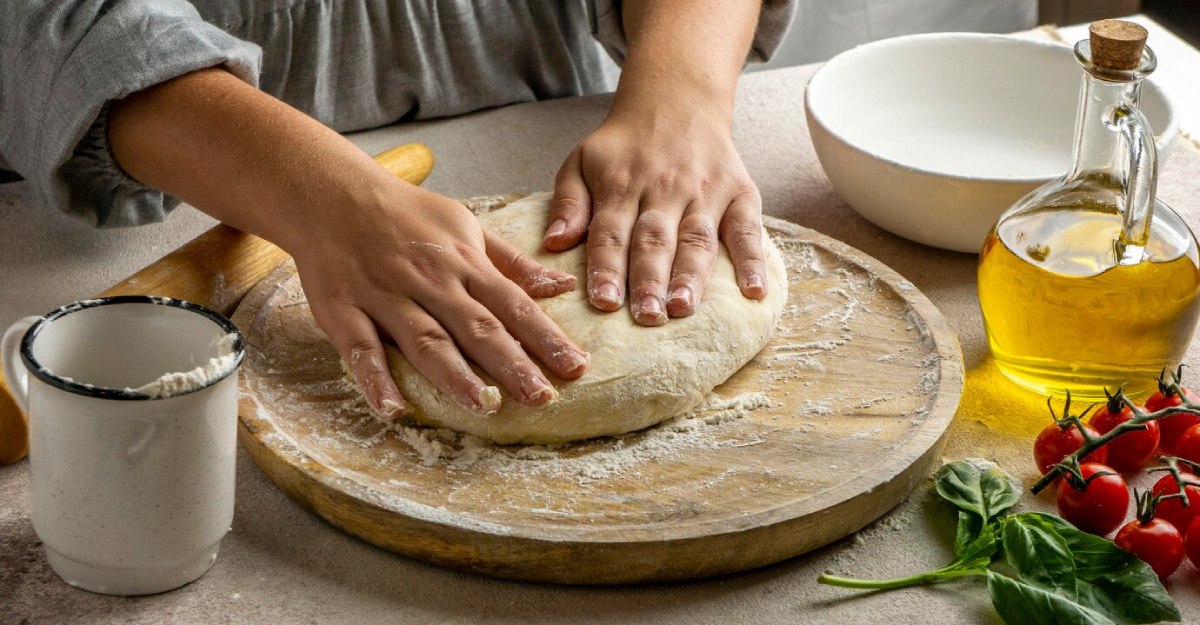 Hands kneading dough on a wooden board, surrounded by ingredients: flour, olive oil, cherry tomatoes, and fresh basil.