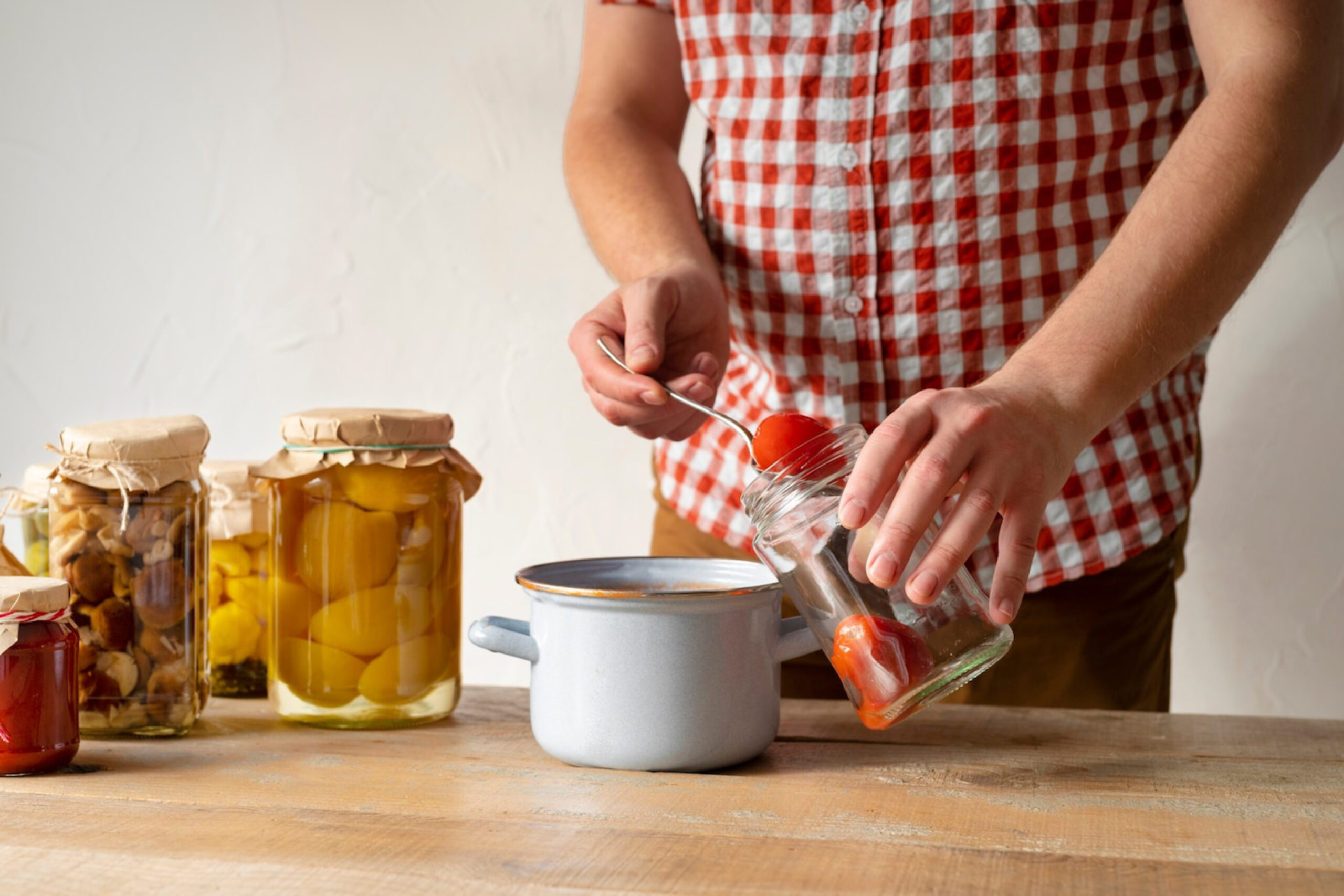 A person in a checked shirt adds tomatoes from a jar into a pot, surrounded by other jars of preserved foods on a wooden table.