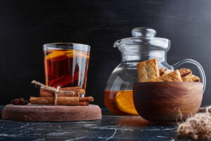 A glass of spiced tea with orange slices, cinnamon sticks, and a wooden bowl of crackers beside a glass pitcher on a dark background.