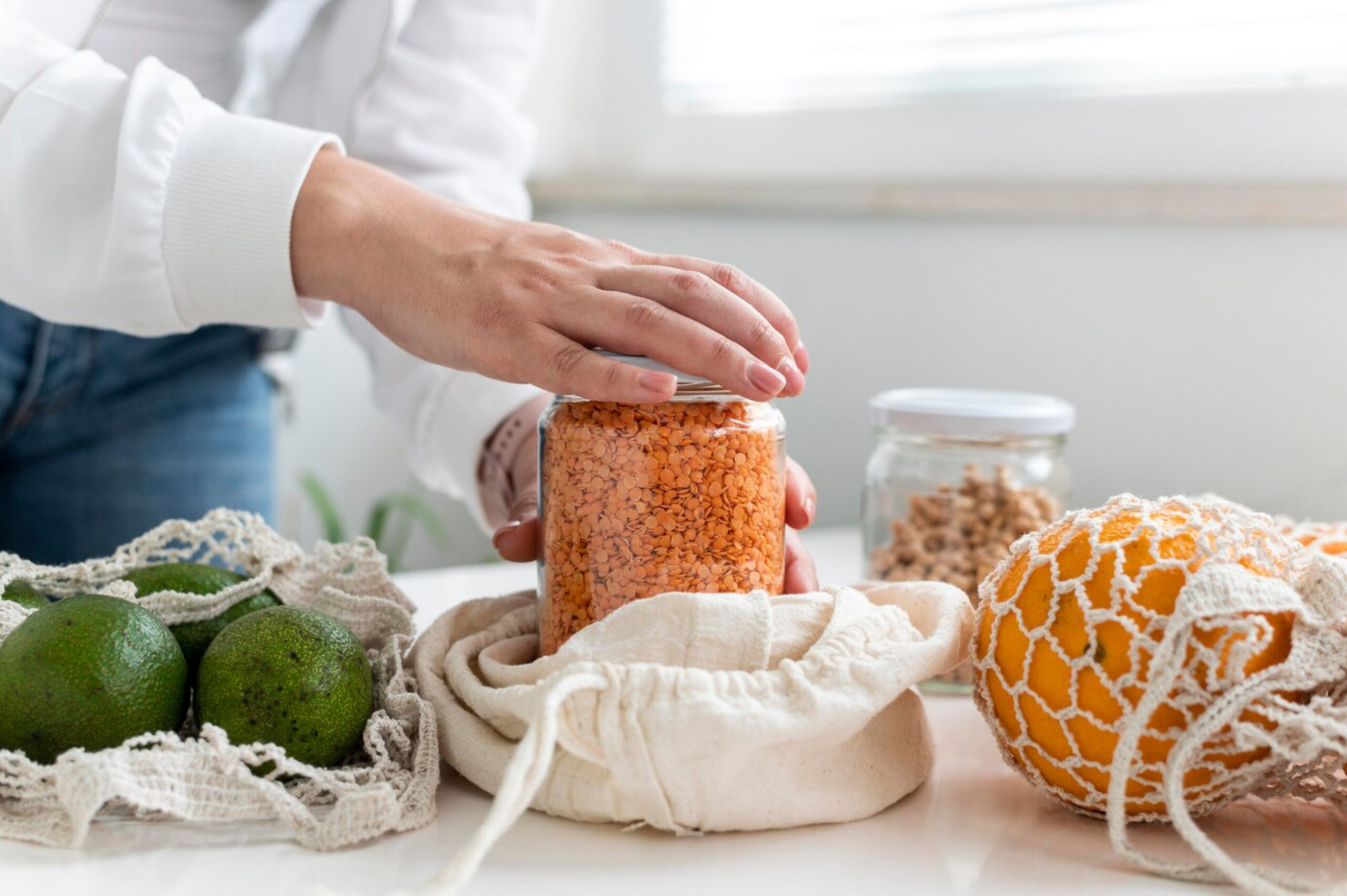 A hand reaches for a jar of orange lentils, surrounded by green avocados and an orange, all placed on a light countertop.