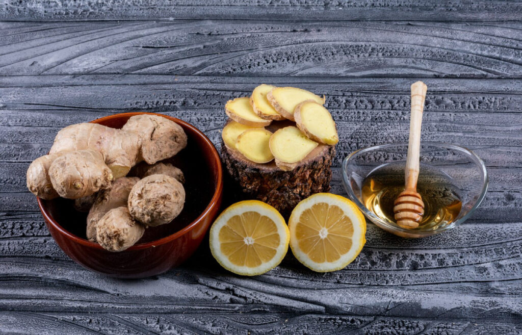 Fresh ginger roots, sliced ginger on a log, honey in a bowl with a dipper, and halved lemons on a textured gray background.