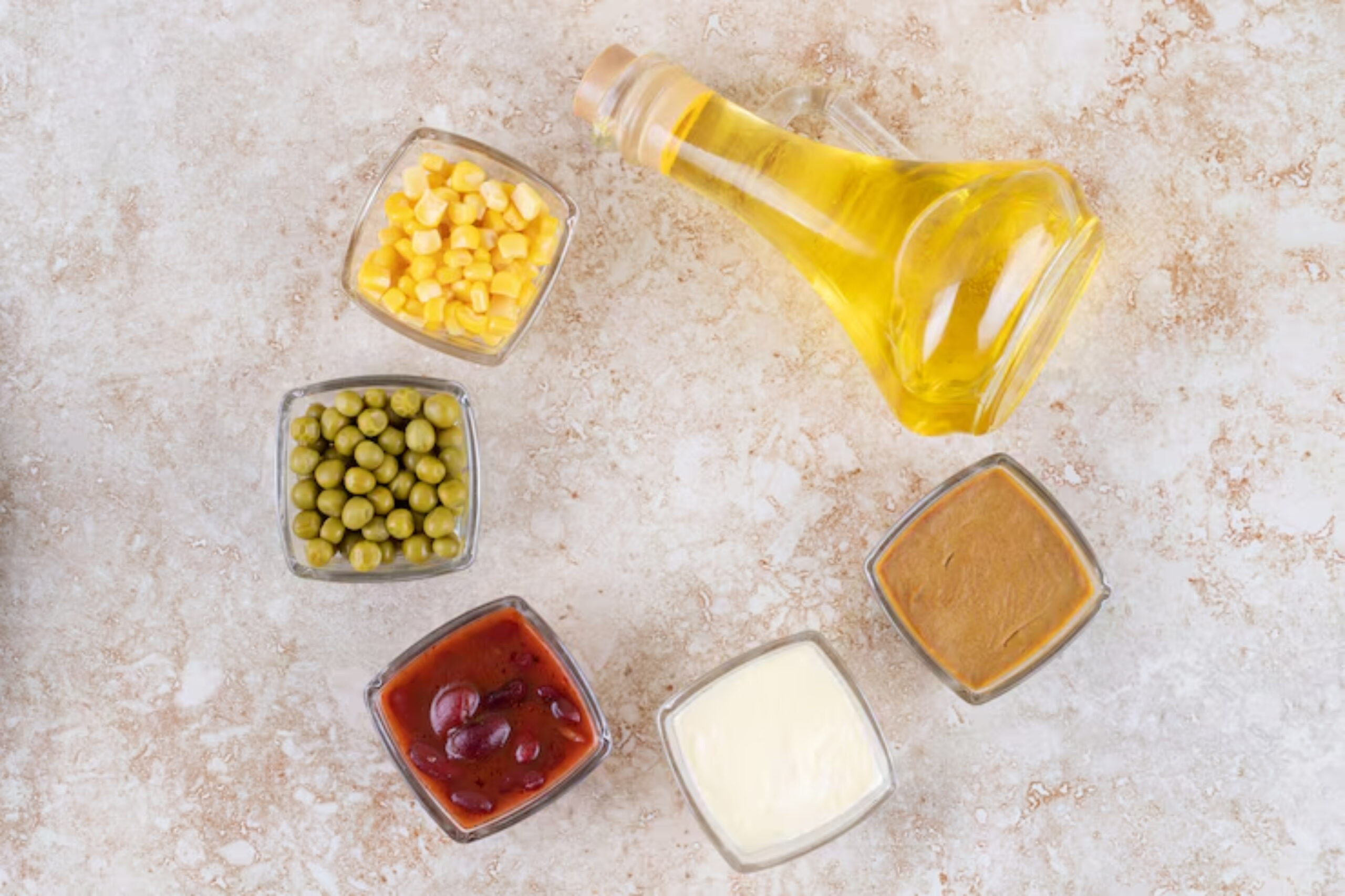 An overhead view of assorted ingredients in glass bowls: corn, peas, kidney beans, sauce, mustard, cream, and a bottle of oil.