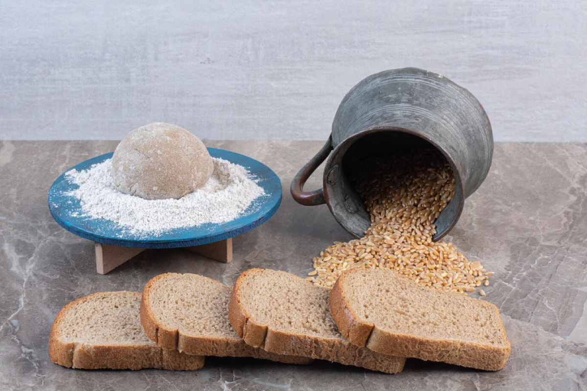 A display of whole grain bread slices, flour, dough ball on a blue plate, and spilled grains from a vintage metal container.