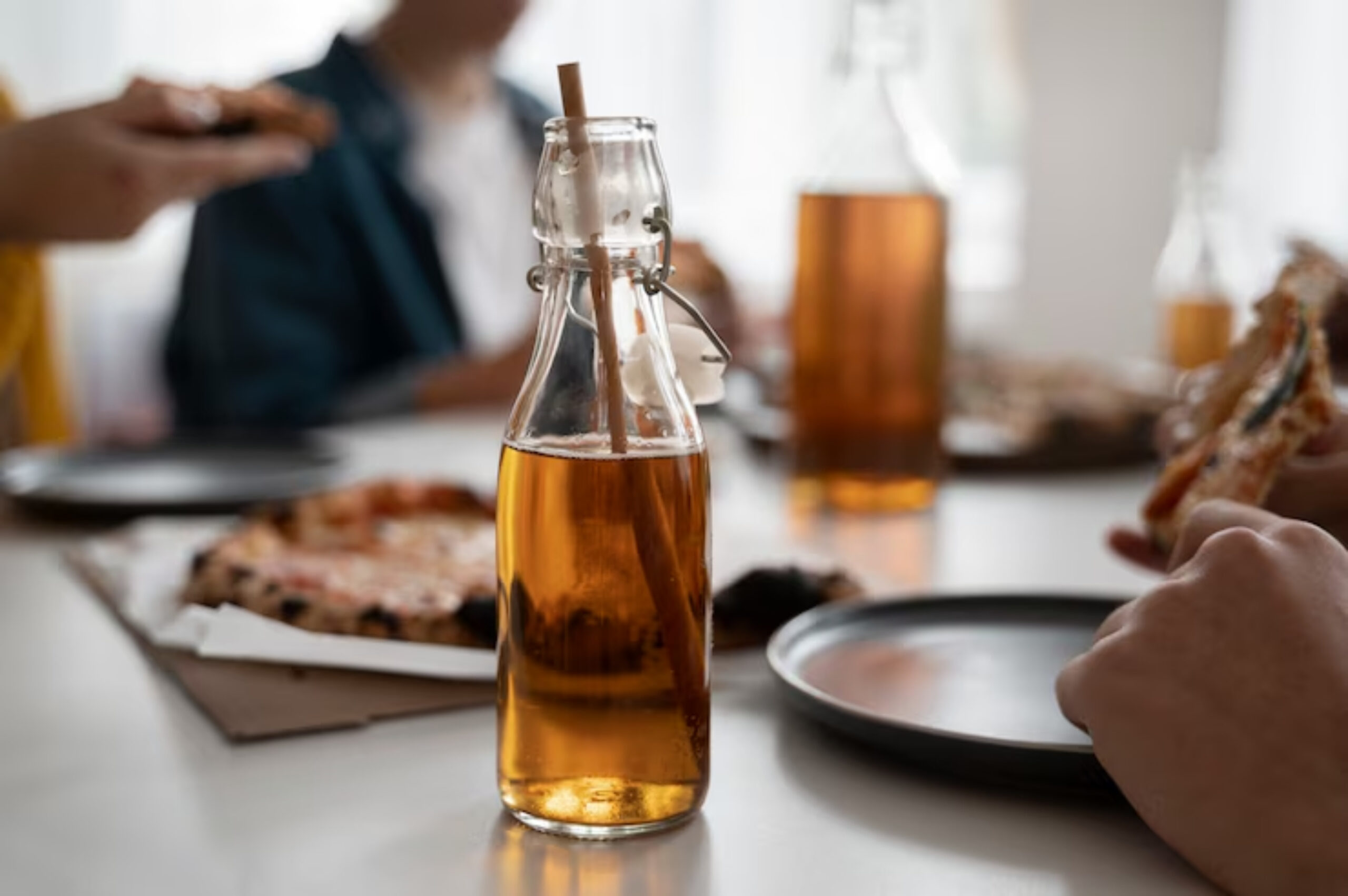 A glass bottle with a straw sits on a table surrounded by friends enjoying pizza and drinks in a casual dining setting.