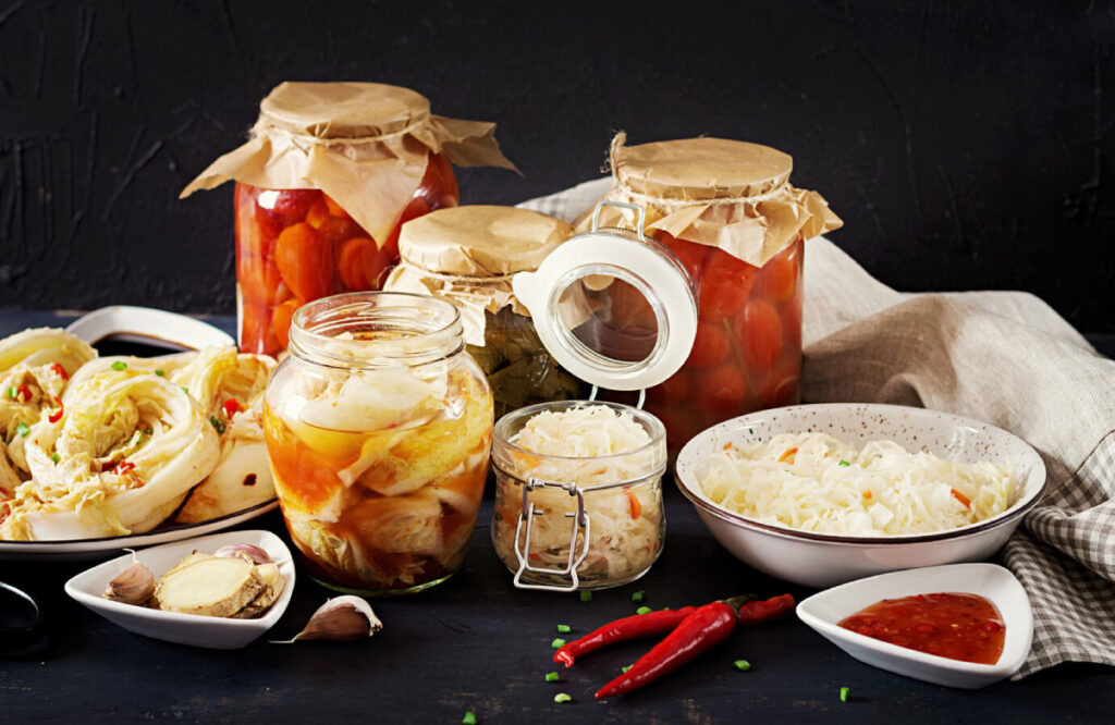 Assorted jars of pickled vegetables, sauerkraut, garlic, ginger, and chili peppers on a dark background, showcasing homemade fermentation.