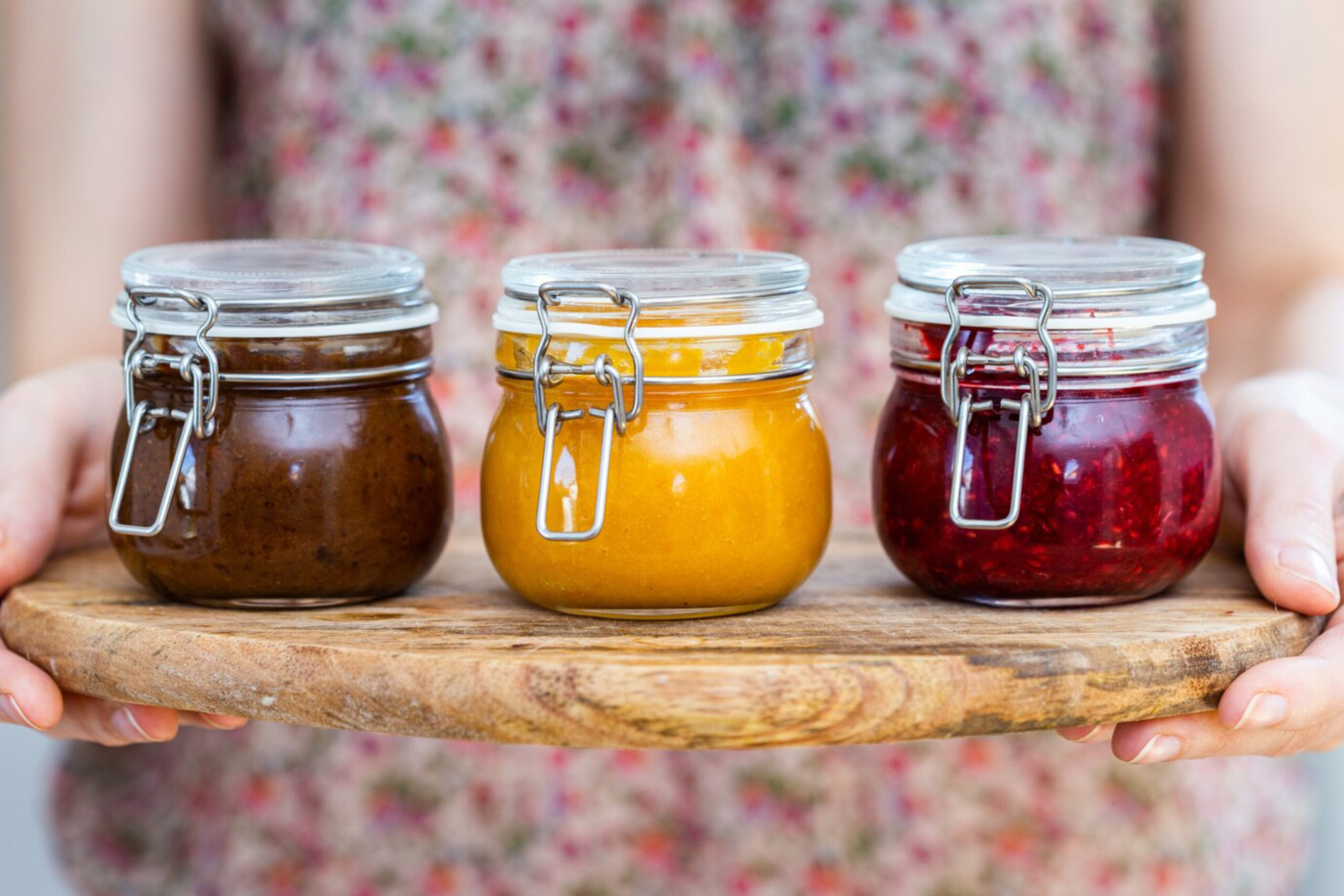 Three glass jars filled with brown sauce, yellow mustard, and red jam, displayed on a wooden platter, held by a person.