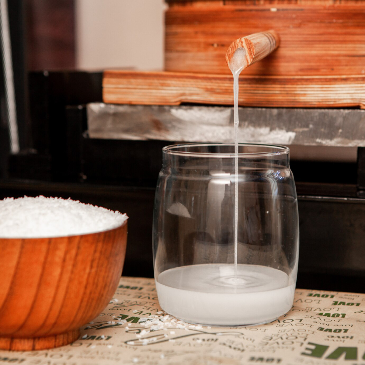 A clear glass jar fills with liquid pouring from a wooden spout, beside a bowl of coarse white salt on a patterned surface.