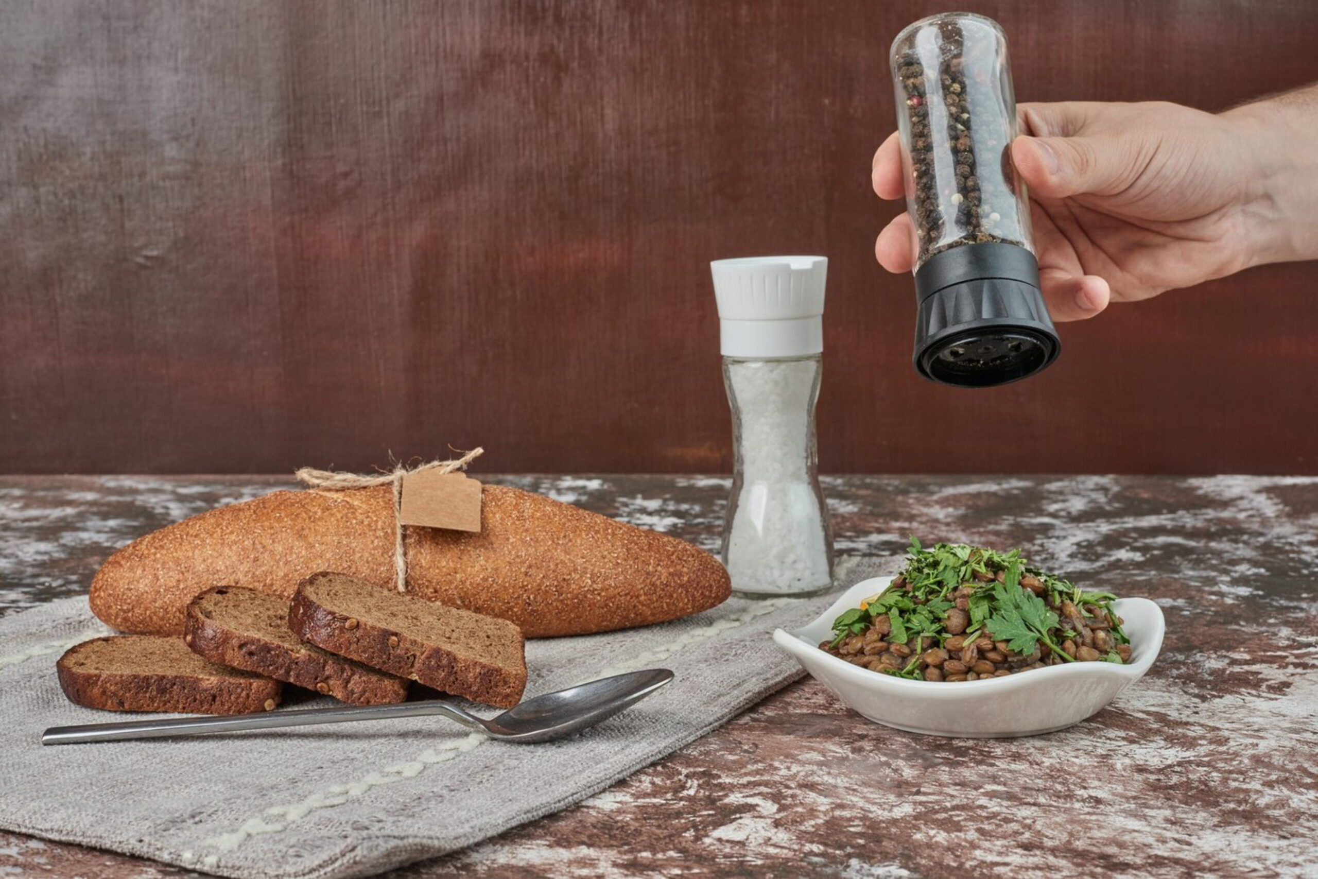 A hand holds a pepper grinder over a bowl of lentils, fresh herbs, next to sliced bread and a salt shaker on a textured table.