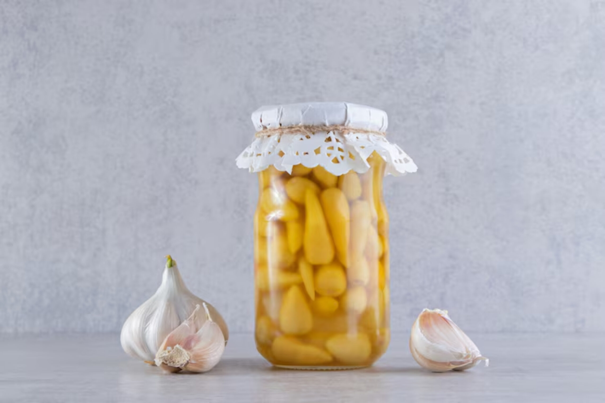 A jar filled with pickled garlic cloves, adorned with a lace paper cover, alongside fresh garlic bulbs on a grey surface.