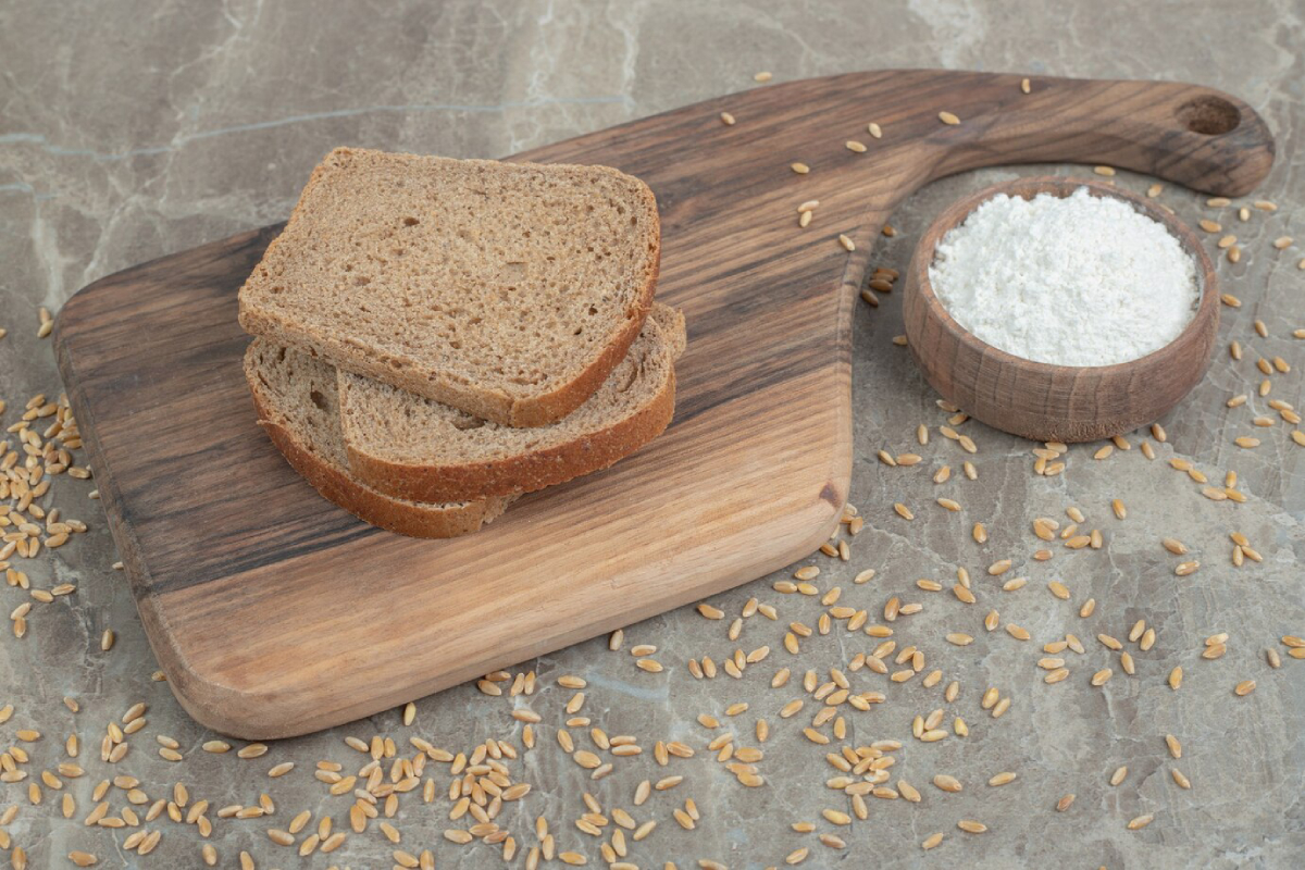 Two slices of brown bread on a wooden cutting board, with a bowl of flour and scattered grains nearby.