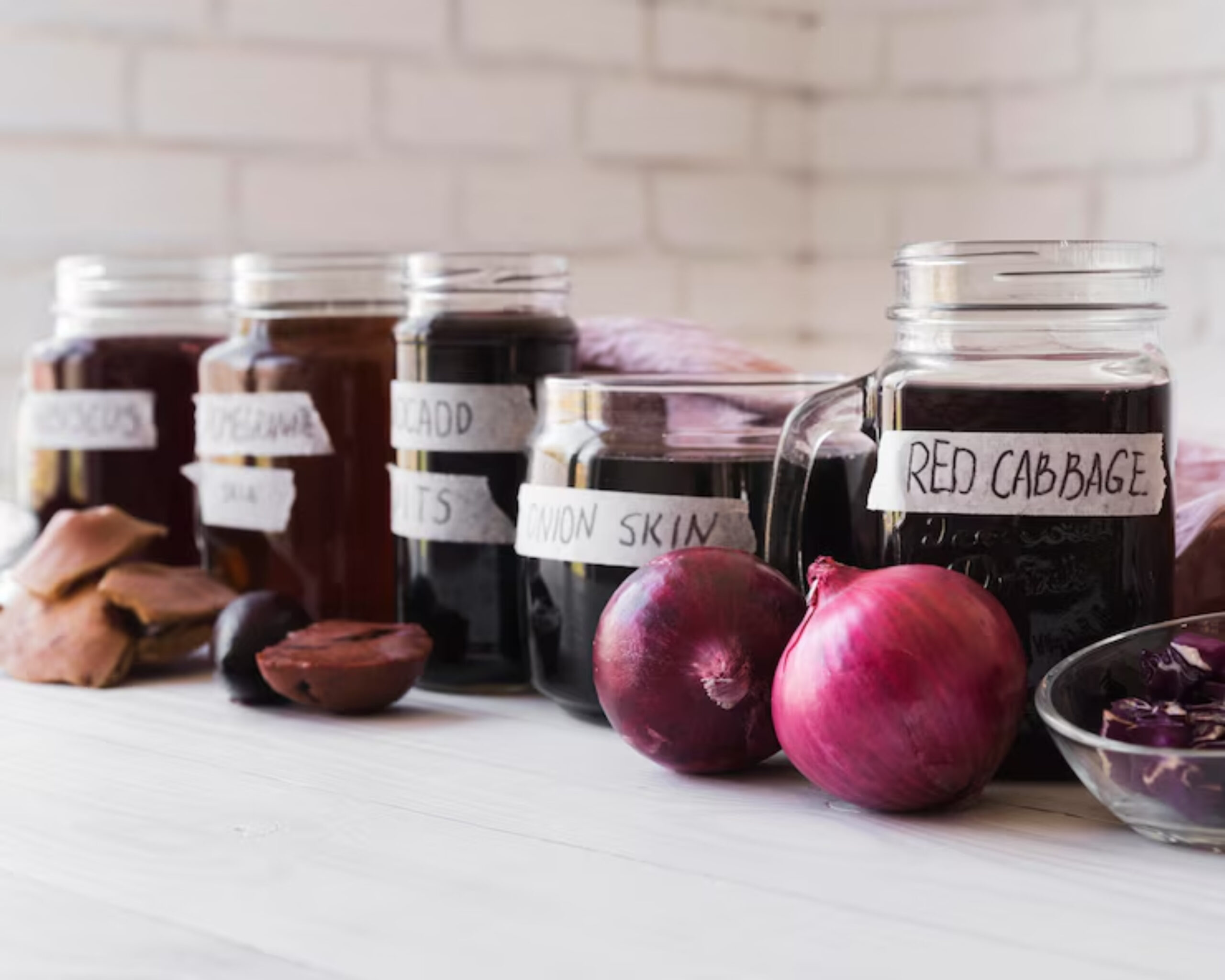Jars of natural dyes labeled with ingredients like hibiscus, avocado pits, and red cabbage, alongside fresh onions and plant materials.