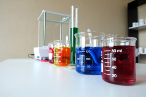 A laboratory countertop displays colorful beakers of liquids: red, blue, yellow, green, and orange, alongside a glass scale and equipment.